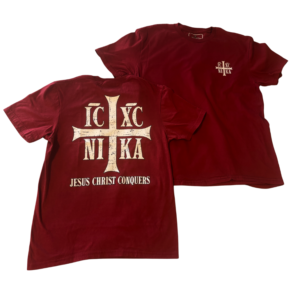 A red short-sleeve t-shirt shown from both the front and back. The back features a large distressed white cross with the Greek letters 'IC XC' and 'NI KA' crossed over it, with 'JESUS CHRIST CONQUERS' printed below.