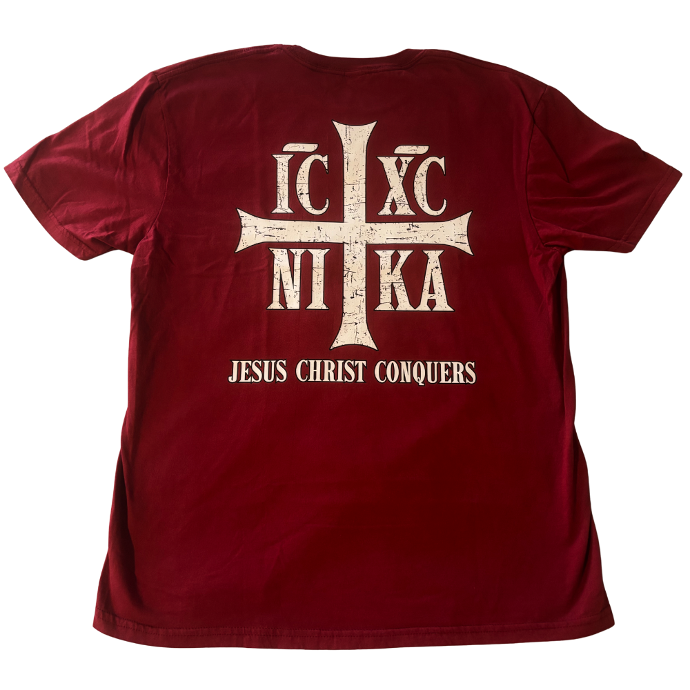 A cardinal red short-sleeve t-shirt shown from the back, featuring a large cream-colored cross with Greek letters 'IC', 'XC', and 'NIKA' and the text 'JESUS CHRIST CONQUERS' printed below it.